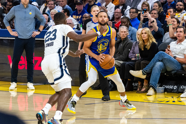 A Golden State Warriors player with the ball is guarded closely by a defender wearing jersey number 23 during a basketball game, with spectators watching.